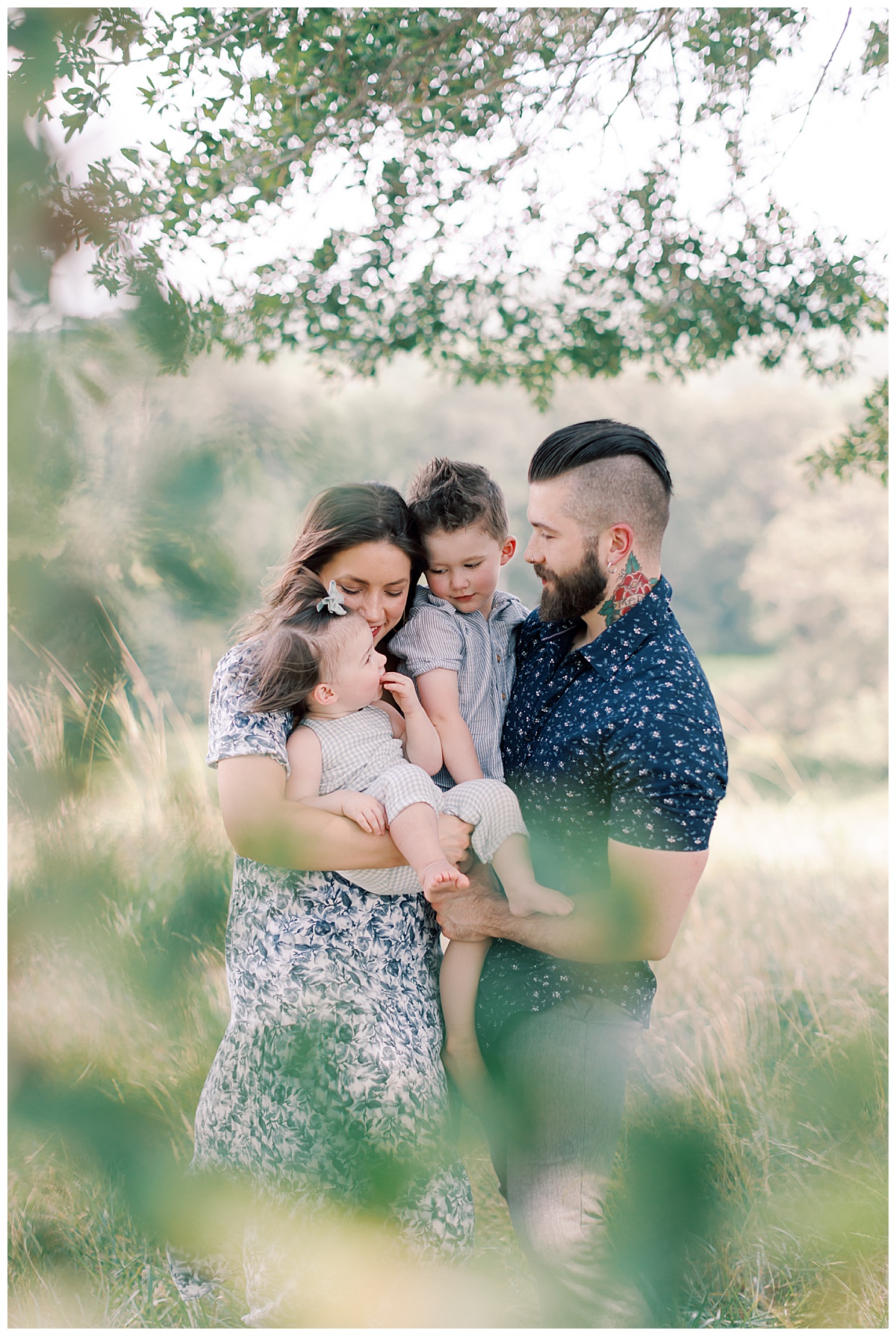 Parents holding their two young daughters together in a sunlit field.