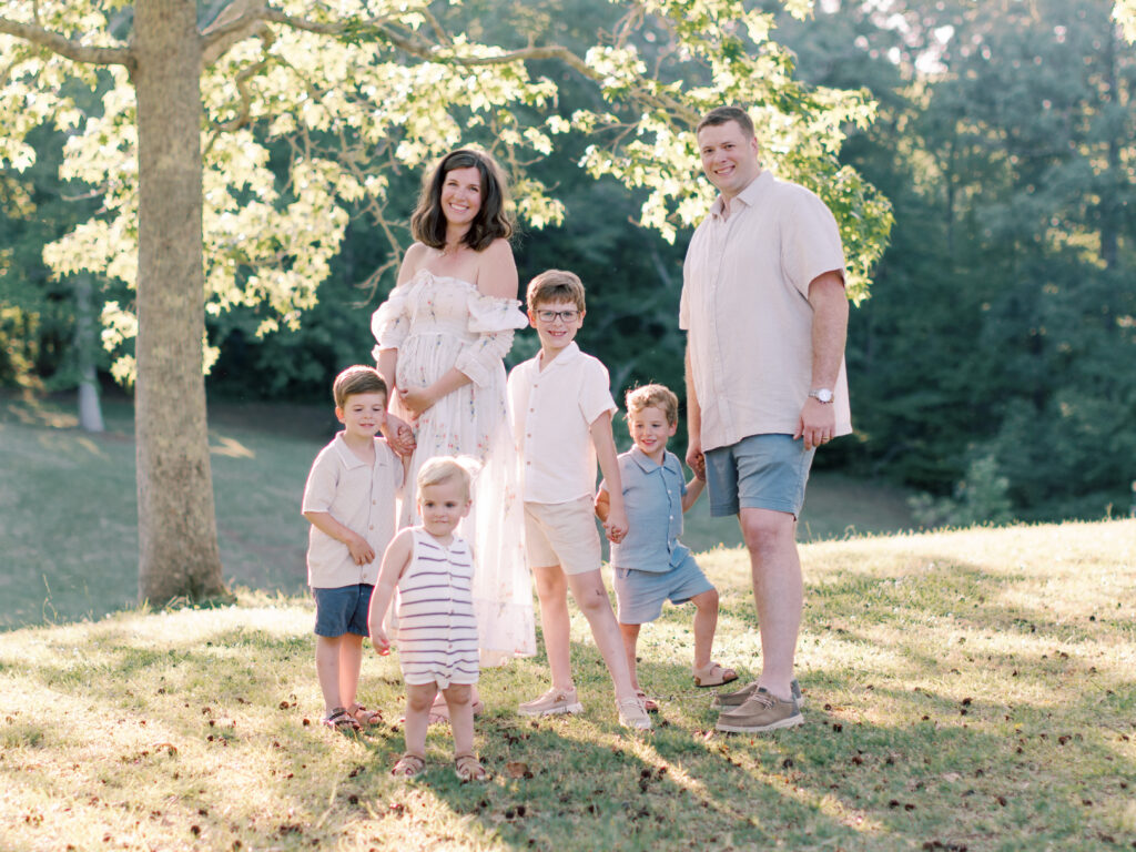 Expectant parents standing with their four young sons beneath a sunlit tree.