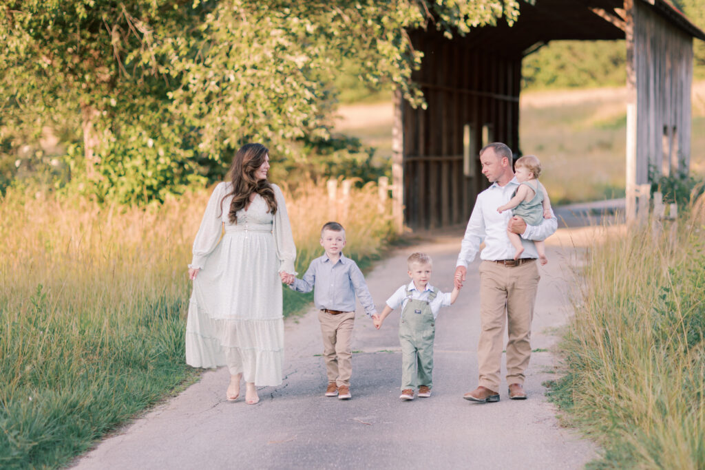 Parents walking hand in hand with their three young children near a covered bridge.