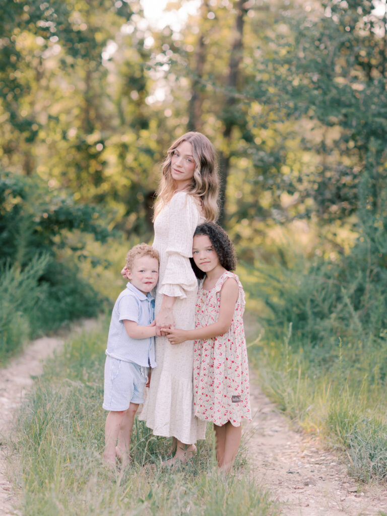 Mother standing on a wooded path as her young son and daughter hug her and lean against her.