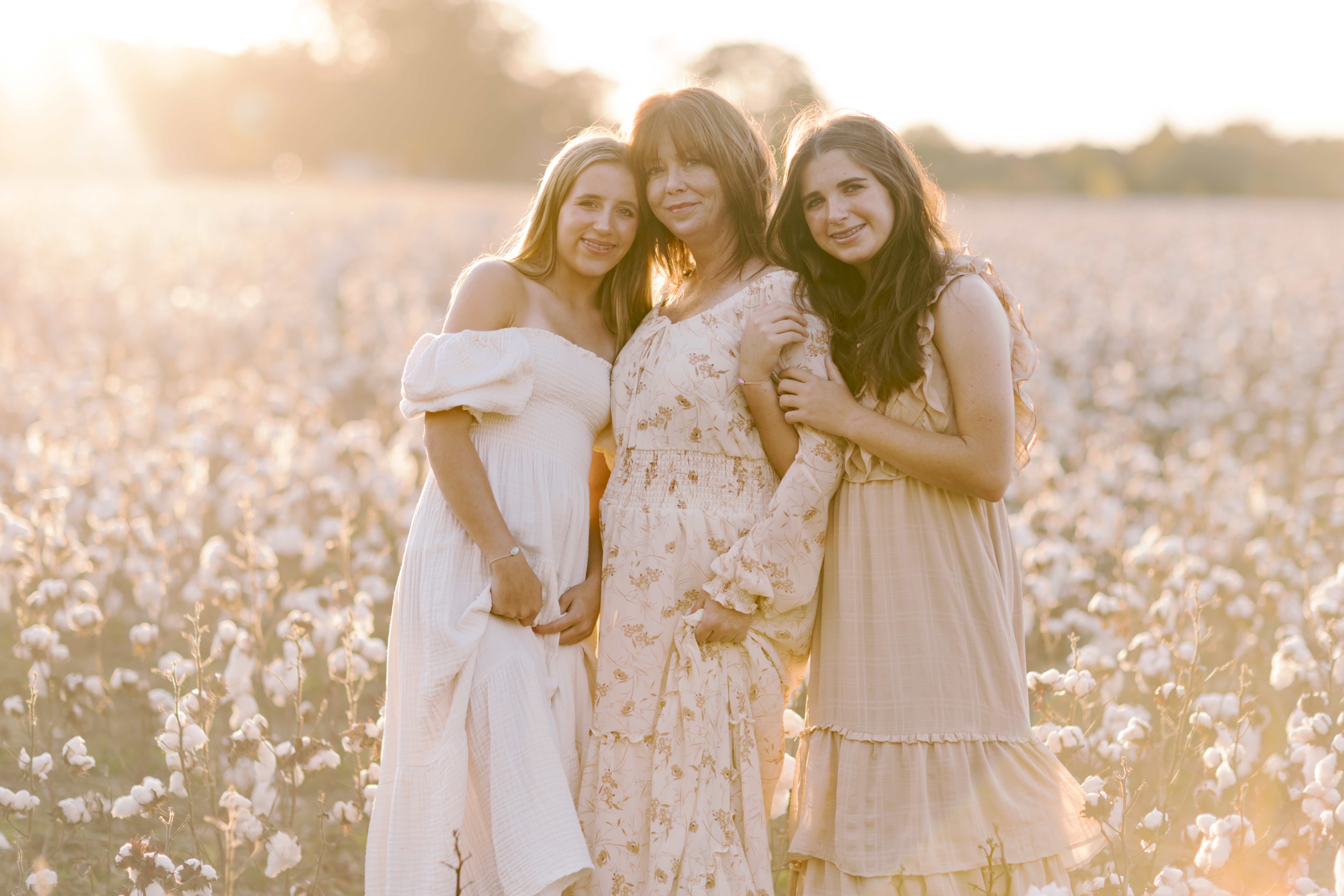 Mother standing closely between her two teenage daughters in a sunlit cotton field.